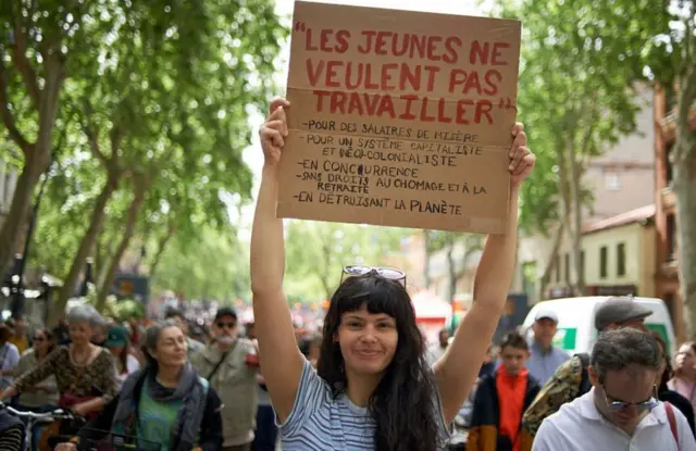 امرأة شابة تحمل لافتة مكتوب عليها: "الشباب لا يرغبون في العمل مقابل أجر زهيد، خاصة في ظل نظام رأسمالي واستعماري جديد يحرمهم من حقوق مثل إعانات البطالة والمعاشات التقاعدية، بينما يستمر في تدمير الأرض".