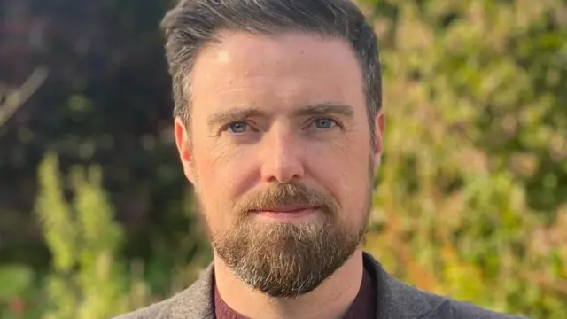 Dr John Paul Loughrey - a man with short dark hair, a beard and moustache looks at the camera smiling. He is wearing a grey suit jacket and red shirt, standing outside with tree's behind him out of focus.
