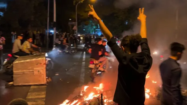 A man stands with his back to camera holding up victory signs at a protest at night