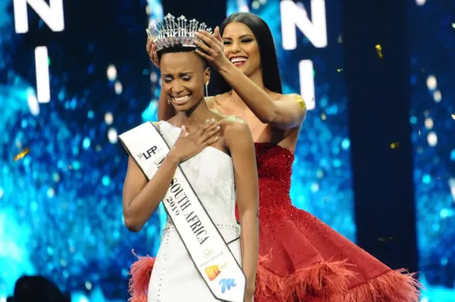 Zozibini Tunzi sourit et serre sa poitrine alors que le diadème du vainqueur est posé sur sa tête.