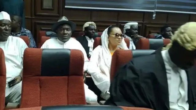 Sịnetọ Natasha yi uwe ọcha nọrọ n'ime ụlọikpe, ebe ụmụnwoke abụọ yikwa uwe ọcha nọ ya n'akụkụ.