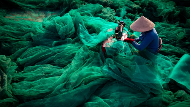 Una mujer sentada frente a su máquina de coser remienda una enorme red de pesca que ocupa el resto de la imagen