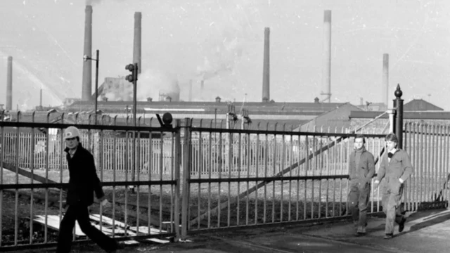 Una foto en blanco y negro de la acería de Corby. Es una enorme fábrica con varias chimeneas. Se ve a lo lejos, tras una valla. Un hombre con casco mira a la cámara mientras camina frente a la valla. Otros dos hombres con monos pasan a poca distancia detrás de él.