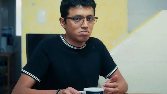 Julian is wearing a navy and white t-shirt and is holding a cup of tea - he has glasses and dark hair 