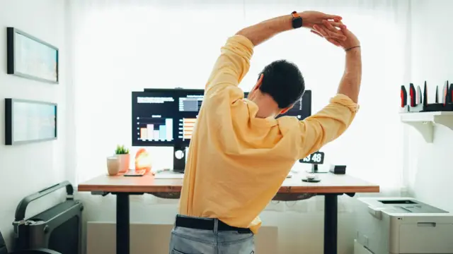 One man wey wear yellow shirt and jeans, from di back. E raise im hands up, hands dey join togeda, and im dey lean to im side as e dey stretch im back.