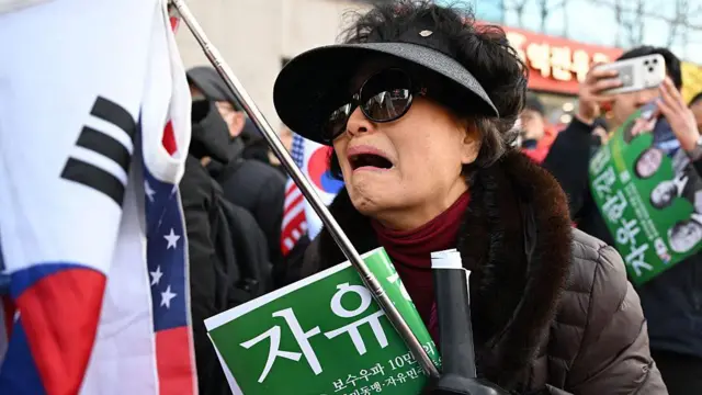 یوه مېرمنه چې تورې عینکې لري بیرغ یې لاس کې دی او ژاړي.