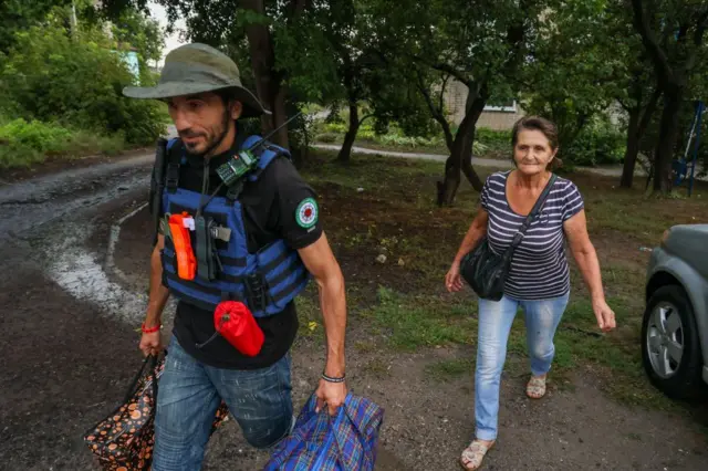 Волонтер у бронежилеті й капелюсі прямує дорогою, несучи дві великі сумки. Поруч іде жінка з ручною сумкою. Дорога ґрунтова, навколо — дерева та житлові будинки. 