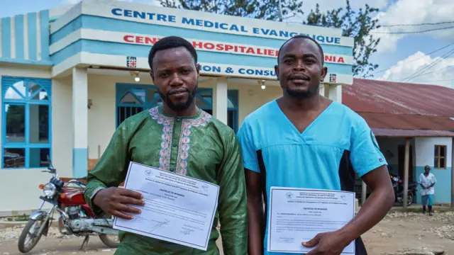 Tagirabo est vêtu de vert et Bulambula de turquoise. Ils tiennent chacun un certificat blanc qu'ils ont reçu pour leur rétablissement et se tiennent devant un centre médical