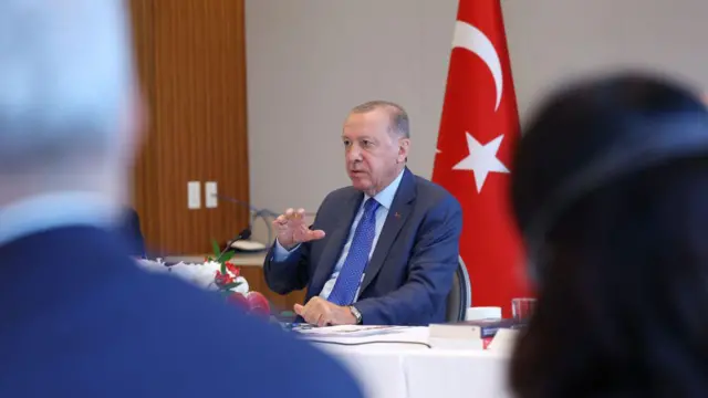 cumhurbaşkanı recep tayyip erdoğan iki kişinin arasından görülüyor. bir masada oturmuş, arkasında türk bayrağı ve önünde mikrofonla konuşma yapıyor
