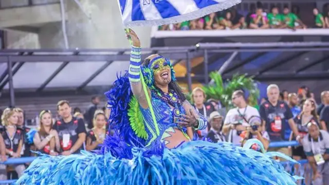 Lucinha Nobre segura bandeira enquanto desfila na Sapucaíaposta na betfair2020, com fantasia azul e verde cheiaaposta na betfairplumas