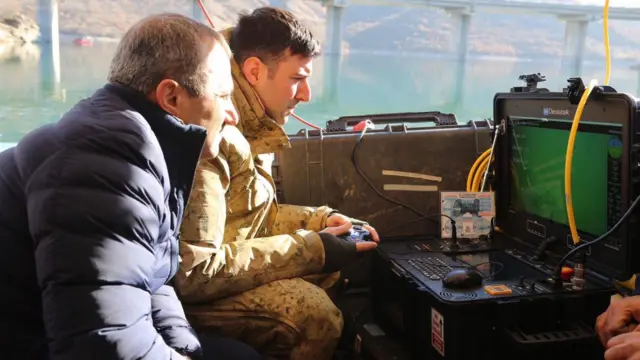 Bir asker ve sivil yetkili bir ekrana bakarak deniz altındaki yapılan aramaya ilişkin görüntüleri izliyor. Arkada bir köprü var. 