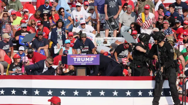 Imagem do palco, decorado com as cores e estrelas da bandeira dos Estados Unidos, com Trump no meio, abaixado e rodeadoroleta roubaseguranças. Ao fundo, diversos apoiadores abaixados. No primeiro plano, um agenteroleta roubasegurança com uma arma.