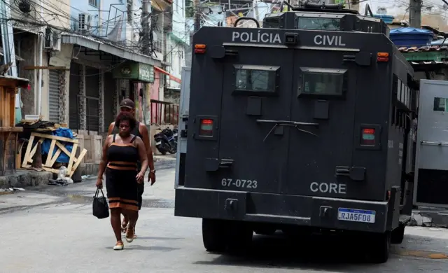 Pessoas passagem ao lado de carro da polícia em comunidade do Rio alvo de operação em 28 de outubro de 2025