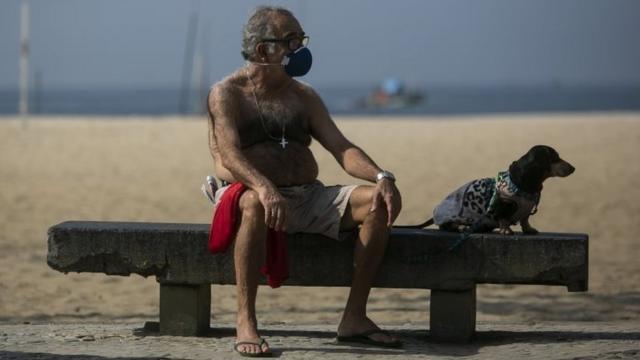 A man wearing a mask sits on a bench with his dog on Rio de Janeiro's Copacabana beach, Brazil. Photo: 17 May 2020