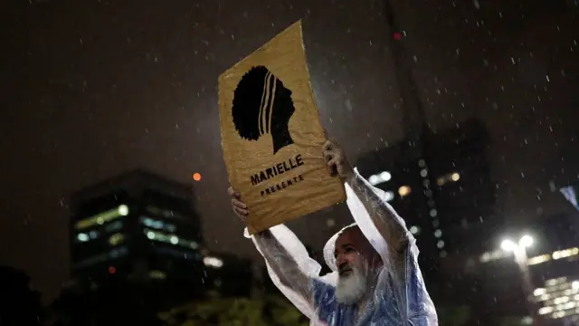 Sob chuva e à noite, homem segura placa com desenho de Marielle e dizeres: 'Marielle presente'
