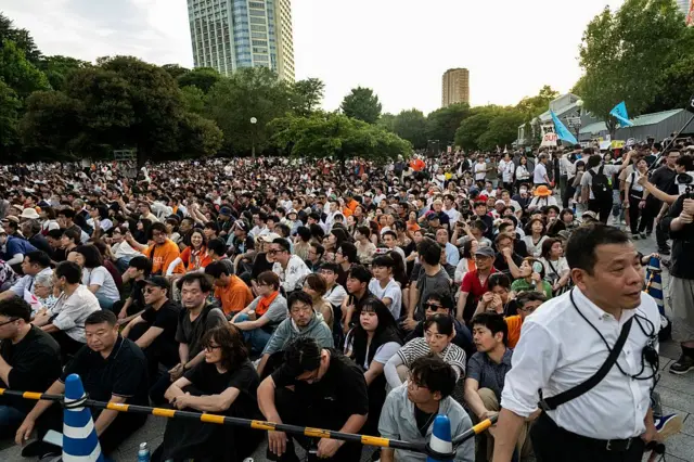 Множество людей стоит и сидит на земле на улице, у некоторых флаги