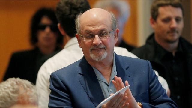 Salman Rushdie olha para a câmera em primeiro plano, com pessoas ao fundo