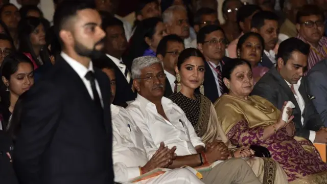 ভারতের রাষ্ট্রপতির কাছ থেকে পুরষ্কার গ্রহণের এক অনুষ্ঠানে ভিরাট কোহলি। পেছনে তার স্ত্রী আনুস্কা শর্মা।