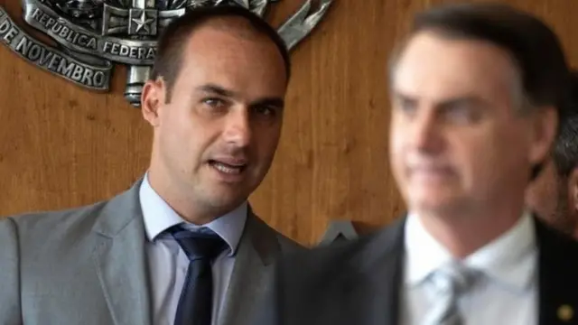 Eduardo Bolsonaro e Jair Bolsonaro, desfocado, está à frente do filho