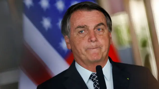 O presidente Jair Bolsonaro aperta os lábios; bandeira dos Estados Unidos ao fundo