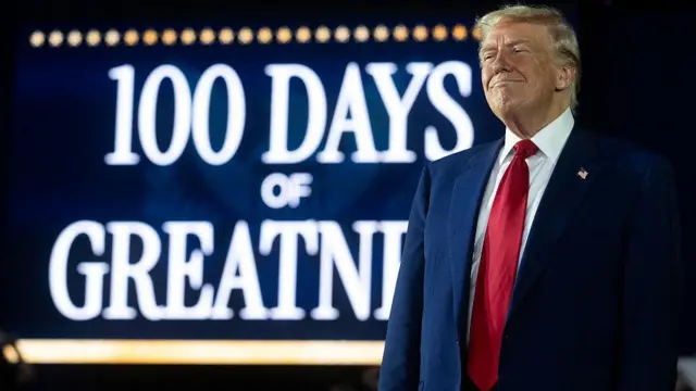 İlginç bir gülüş takınan Trump'ın arkasında "100 Günlük Mükemmellik" yazan bir tabela var.