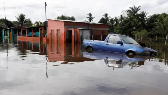 Carro em meio a água, com casas ao fundo