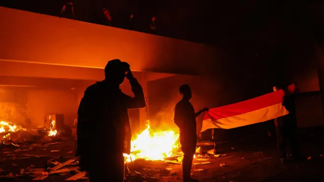 Pengunjuk rasa membentangkan umbul-umbul merah putih di depan gedung DPRD Sulawesi Selatan yang dilalap api saat aksi 29 Agustus di Makassar, Sulawesi Selatan, Sabtu (30/8/2025) dini hari. 