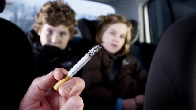 No primeiro plano, uma mão segura um cigarro aceso. No segundo plano, duas crianças - um menino e uma menina - no banco de trás de um carro