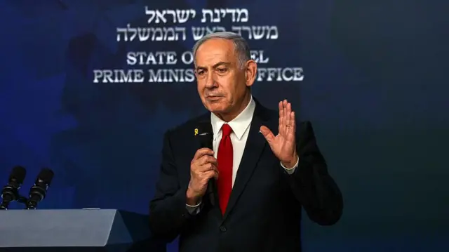 رئيس الوزراء الإسرائيلي، بنيامين نتنياهو، يحمل ميكرفوناً مرتدياً سترة سوداء وقميصاً أبيض وربطة عنق سوداء.