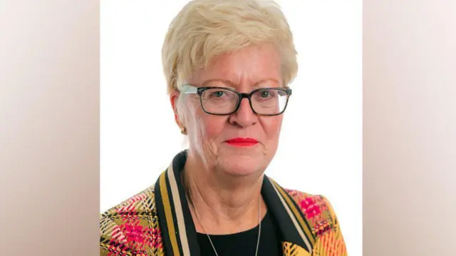 Helen Pitcher has short blonde hair and a red green and yellow tartan jacket on with a black collar with yellow stripes on it. She is wearing glasses. 