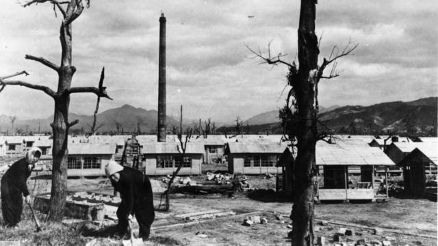 Dos mujeres construyen casas precarias de emergencia tras la bomba atómica. Junto a ellas se ven los troncos carbonizados de dos árboles.