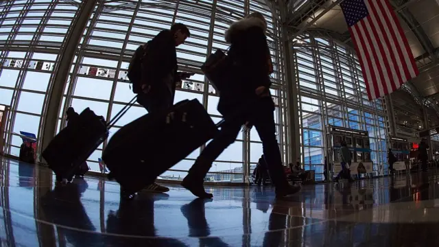 Duas pessoas caminham carregando malas em corredor de aeroporto, com bandeira dos EUA pendurada
