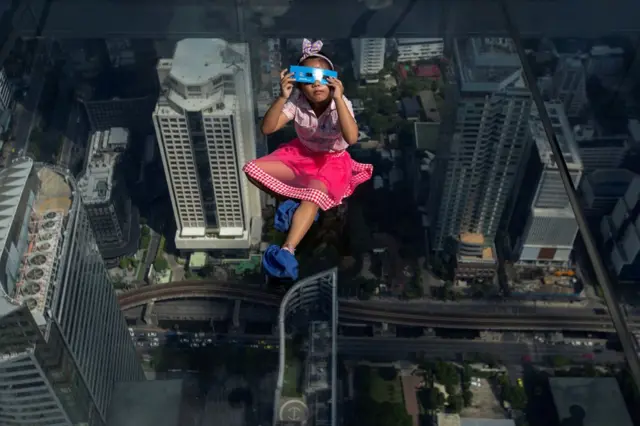 A girl looks through an eclipse viewer on an elevated, transparent platform, with buildings and roads in the distance below her