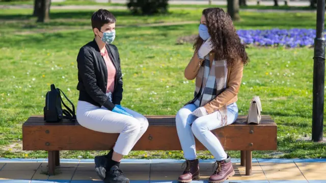 Dos mujeres con mascarilla sentadas en una banca en un parque.