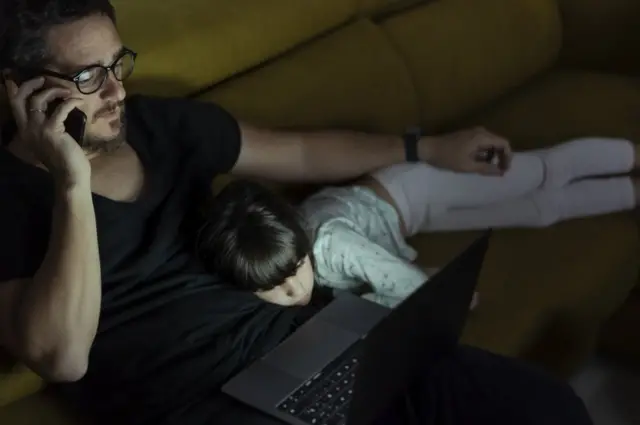 Un hombre teletrabajando hasta tarde con su niña acostada sobre sus piernas.