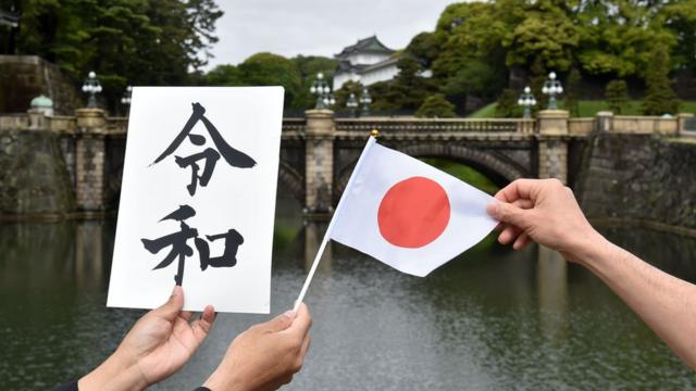 People hold a flag of Japan and a board bearing new era name "Reiwa" near the Double Bridge at the Imperial Palace in Tokyo on May 1,
