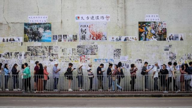 People queue to cast their vote in front of a "Lennon Wall" adorned with tattered posters