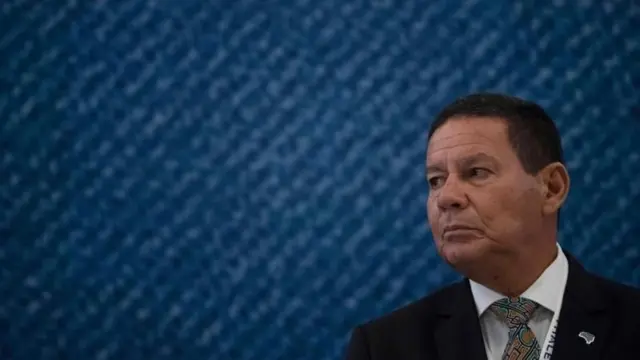 Mourão olhando para o lado, com olhar sério, em frente a painel azul