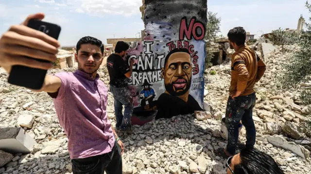 A friend of the muralist Aziz Asmar takes a selfie as the artist works on his Floyd memorial in Idlib, Syria