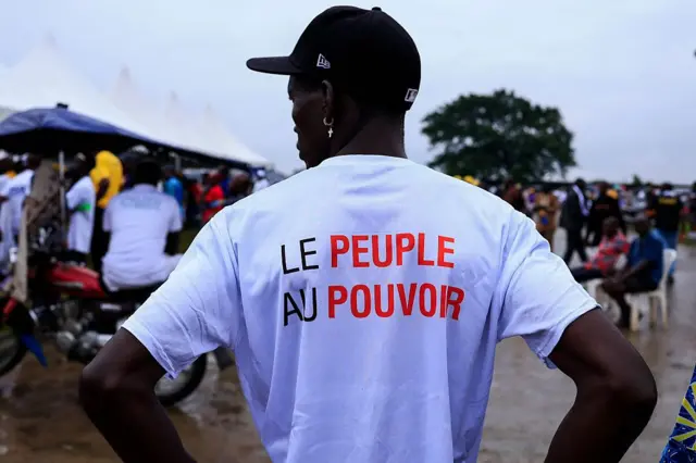 Un homme portant un t-shirt blanc sur lequel est écrit "Le peuple au pouvoir", avec en arrière-plan une foule de personnes.