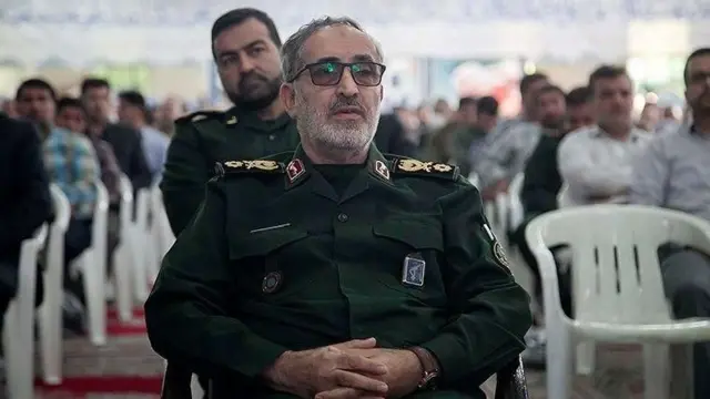 علی شادمانی در یک جلسه سخنرانی با لباس نظامی روی صندلی نشسته است
