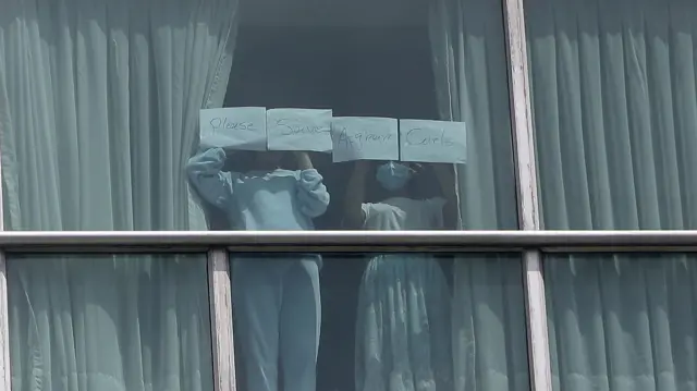 Two young migrants stand at a hotel window with a message asking for help for Afghan girls