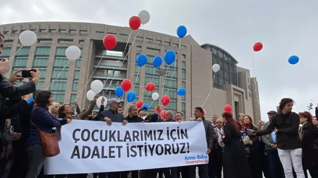 Kalabalık "Çocuklarımız için adalet istiyoruz" yazan bir pankart tutuyor, kırmızı, beyaz, mavi balonlar .