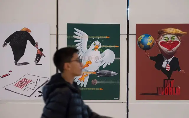 یک پسر رهگذر در تهران که پشت سرش و روی دیوار بیلبوردهایی علیه ترامپ و آمریکا نصب شده است