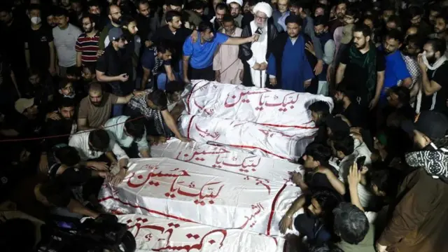 白地に赤でアラビア文字の書かれた布でくるまれた複数の棺の周りを、多くの男性が取り囲んでいる