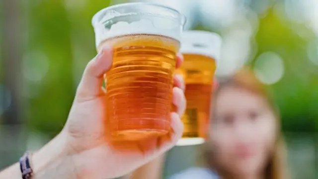 Mãos segurando copos com bebidas, com mulher ao fundo