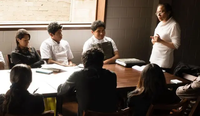 Mujer enseña a unos jóvenes sentados en una mesa.