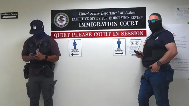 အမေရိကန် လူဝင်မှုကြီးကြပ်ရေးတရားရုံးအတွင်းမှာ ဝတ်စုံပြည့်နဲ့ မတ်တပ်ရပ်နေတဲ့ ICE တပ်ဖွဲ့ဝင် ၂ ဦး 