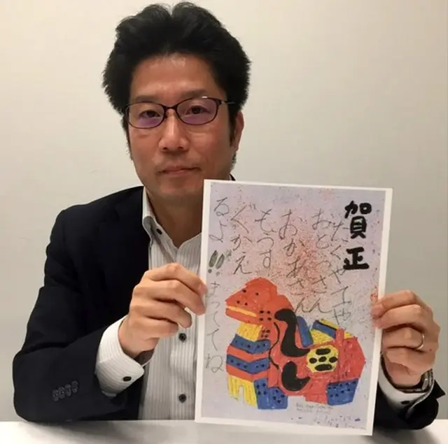 Takuya Yokota, in a black suit, holds up a copy of a postcard featuring a colourful animal