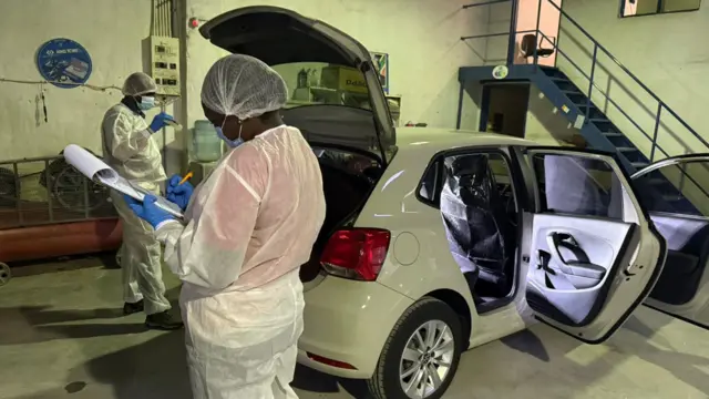 Investigators wearing white dey inspect a white VW Polo vehicle. She dey hold book